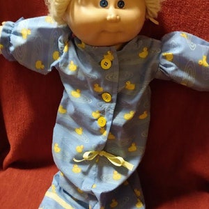 May include: A vintage doll with a blonde yarn wig and blue eyes, wearing blue pajamas. The pajamas feature a pattern of yellow ducks on a water-like design. The outfit has yellow buttons and a yellow ribbon tie at the waist.