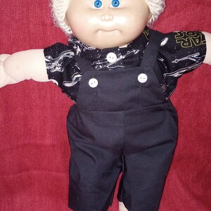 May include: A Cabbage Patch Kid doll with blonde hair wearing a black overall with white buttons and a black and white shirt with a space theme print.