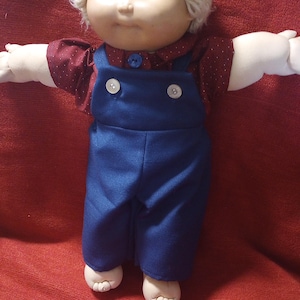 Pode incluir: Uma boneca com rosto redondo, olhos azuis e cabelo curto de lã loira. A boneca veste uma camisa vermelho-escura com pequenos pontos brancos e jardineiras de ganga azul com dois botões brancos e um botão azul na gola.