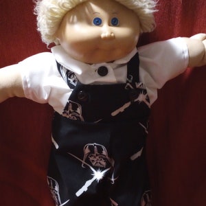 May include: A Cabbage Patch Kid doll wearing a white shirt and black overalls with a repeated pattern of Darth Vader's face. The doll has blonde hair and blue eyes.