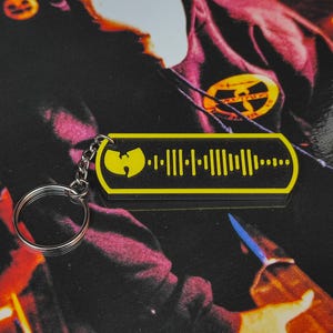 May include: A black and yellow keychain with a music waveform design and a yellow logo. The keychain is attached to a silver key ring. The background is a dark, patterned surface.