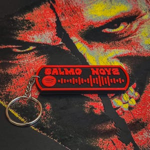 May include: A black and red keychain featuring the text "SALMO NOYZ" and a Spotify code. The keychain has a silver key ring and is set against a dark, textured background with red and yellow accents.