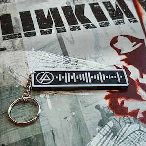 May include: A black and white keychain with a barcode design and a band logo. It has a silver keyring. The background features the word "LINKIN" in black lettering.