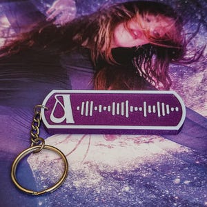 以下が含まれることがあります： 金色のキーリングが付いた、紫と白のキーホルダー。キーホルダーは長方形で、白い縁と紫色の中心があります。中央には白い文字「A」と音波のデザインがあります。