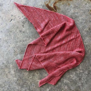 May include: A knitted triangular shawl in a muted red colour. The shawl has a textured stitch pattern and an openwork design. The fabric is draped on a grey surface, showing its shape and texture. The shawl is likely made of wool or a similar fibre.