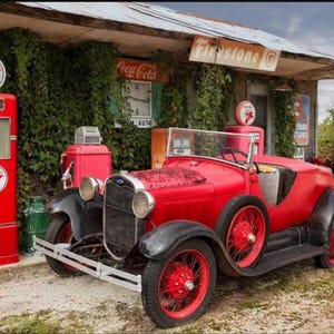 Puede incluir: Un coche rojo antiguo Ford Model A roadster con guardabarros negros y ruedas rojas, estacionado junto a una bomba de gasolina Texaco roja. Un letrero de Coca-Cola y un letrero de Firestone son visibles en el fondo.
