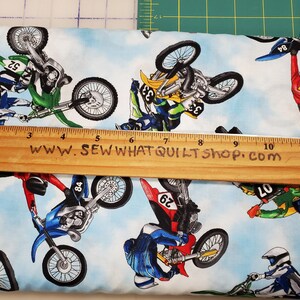 K&ouml;nnte beinhalten: Ein wei&szlig;er Stoff mit blauem Himmel als Hintergrund und einem sich wiederholenden Muster von Motocross-Fahrern, die Stunts auf ihren Motorr&auml;dern ausf&uuml;hren. Die Fahrer tragen bunte Rennkleidung und die Motorr&auml;der sind in verschiedenen Farben, darunter Gr&uuml;n, Blau, Gelb und Rot. Der Stoff tr&auml;gt den Text "www.sewwhatquiltshop.com".