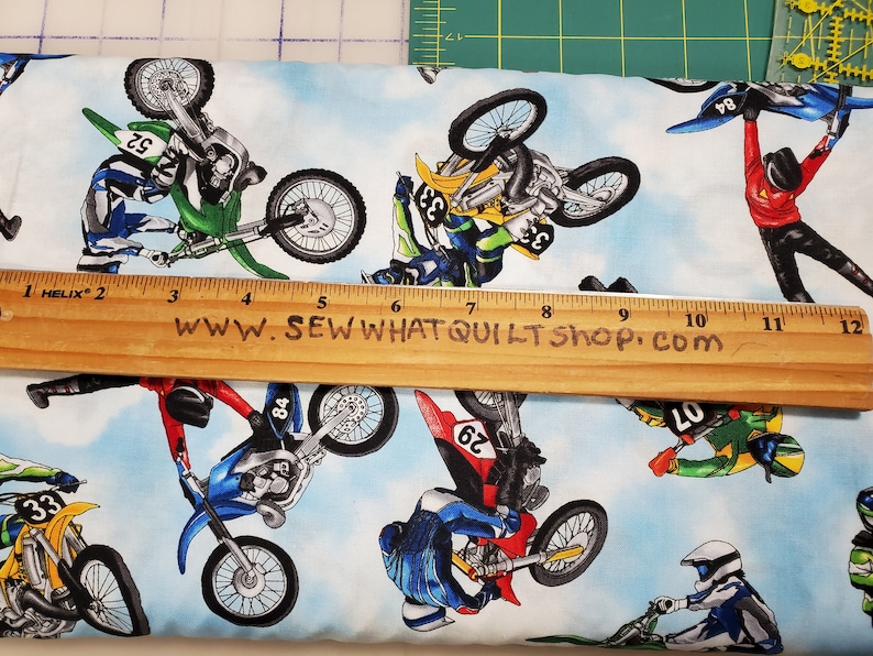 K&ouml;nnte beinhalten: Ein wei&szlig;er Stoff mit blauem Himmel als Hintergrund und einem sich wiederholenden Muster von Motocross-Fahrern, die Stunts auf ihren Motorr&auml;dern ausf&uuml;hren. Die Fahrer tragen bunte Rennkleidung und die Motorr&auml;der sind in verschiedenen Farben, darunter Gr&uuml;n, Blau, Gelb und Rot. Der Stoff tr&auml;gt den Text "www.sewwhatquiltshop.com".