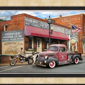 Puede incluir: Una impresión enmarcada que presenta una escena vintage con una camioneta roja estacionada frente a un edificio de ladrillo. El edificio tiene un toldo rojo y letreros que dicen "Frontier Hardware Co. Inc." y "Motorcycle Club Garage". Una bandera estadounidense ondea.