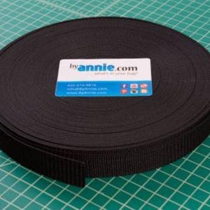 May include: A roll of black webbing material sits on a green cutting mat. A business card with the text "byannie.com" is on top of the roll. The card has contact information and social media icons.