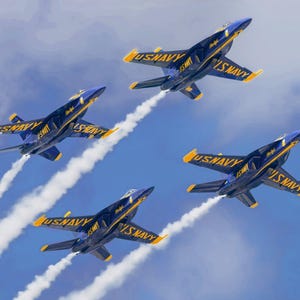 May include: Four blue and yellow US Navy jets in formation against a blue sky. The jets are leaving white contrails. The text "US NAVY" is visible on the side of each jet.
