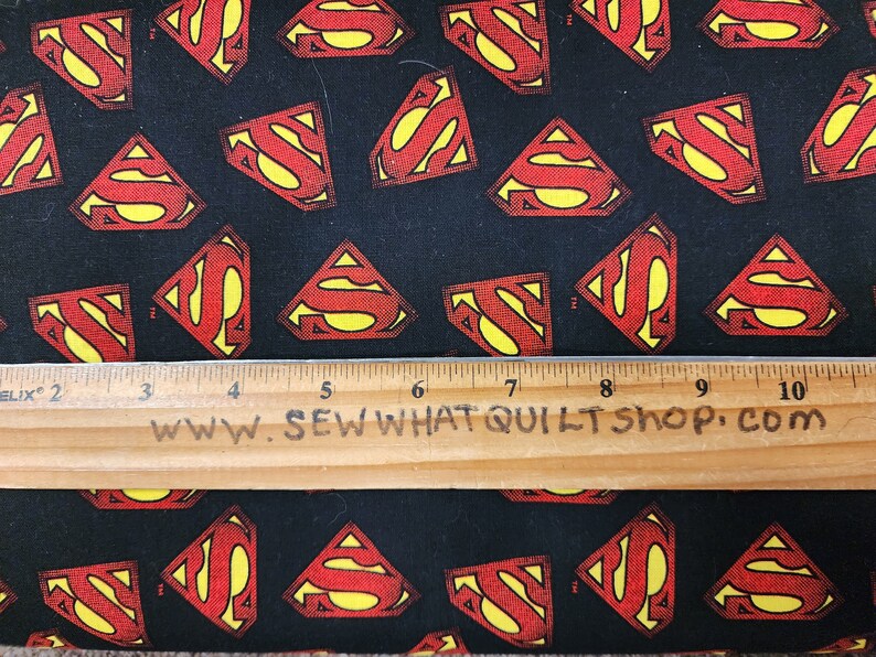 May include: Black fabric with a repeating pattern of red and yellow Superman logos. A wooden ruler with the text "www.sewwhatquiltshop.com" is placed on top of the fabric.