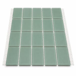 May include: A rectangular sheet of sage green glass subway tiles arranged in a grid pattern. The individual tiles have smooth, glossy surfaces and rounded edges, mounted on a white mesh backing that is visible between each tile row and column.