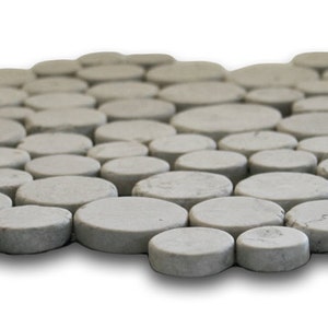 May include: A gray, pebble-like mosaic tile mat. The mat is composed of numerous, irregularly shaped, flat, circular stones. The stones are arranged in a slightly curved pattern, creating a textured surface. The color is a neutral gray, with subtle variations in tone.