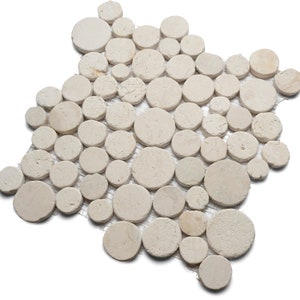 May include: A mosaic tile sheet composed of numerous beige, circular stones of varying sizes. The stones are arranged on a mesh backing, creating a natural, textured surface. The neutral color palette and organic shapes suggest a spa or bathroom theme.