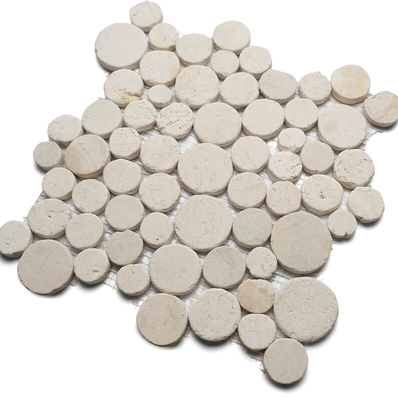 May include: A mosaic tile sheet composed of numerous beige, circular stones of varying sizes. The stones are arranged on a mesh backing, creating a natural, textured surface. The neutral color palette and organic shapes suggest a spa or bathroom theme.