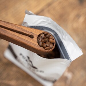 May include: A wooden coffee scoop with a handle, filled with dark brown coffee beans, being poured into a white bag with black text.