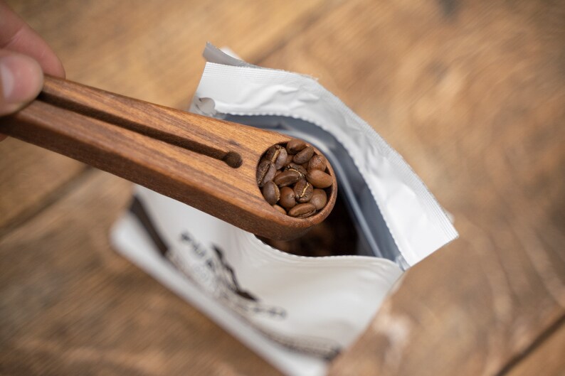 May include: A wooden coffee scoop with a handle, filled with dark brown coffee beans, being poured into a white bag with black text.