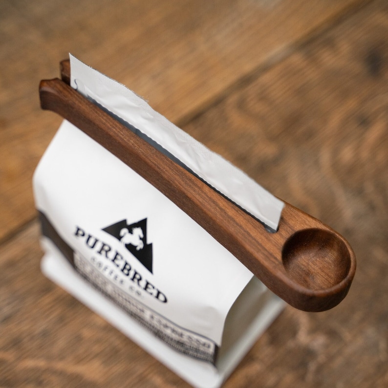 May include: A wooden coffee scoop with a handle attached to a white paper bag with the logo "PUREBRED COFFEE CO."