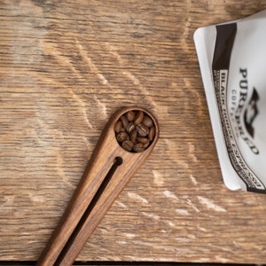 May include: A wooden coffee scoop with a dark brown finish and a small, round compartment filled with coffee beans. The scoop is on a wooden surface.