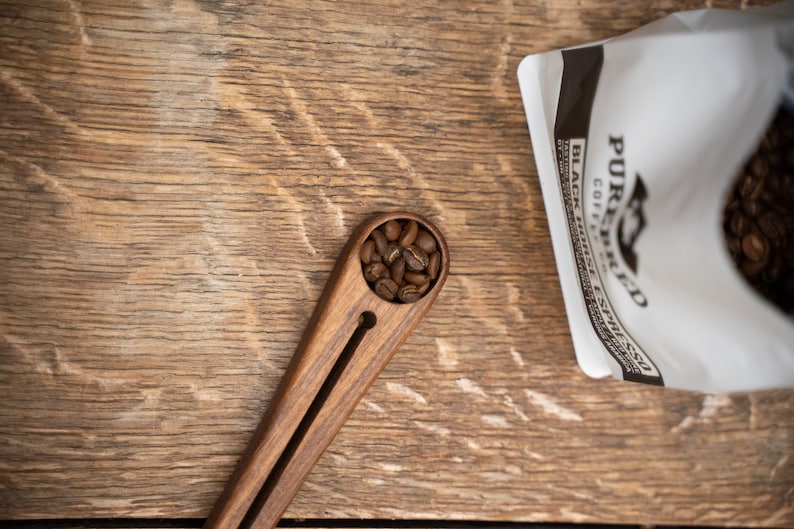 May include: A wooden coffee scoop with a dark brown finish and a small, round compartment filled with coffee beans. The scoop is on a wooden surface.