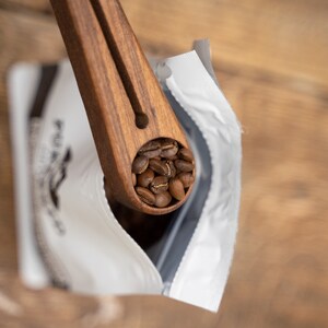 May include: A wooden scoop filled with roasted coffee beans, being poured into a white bag with a black label.