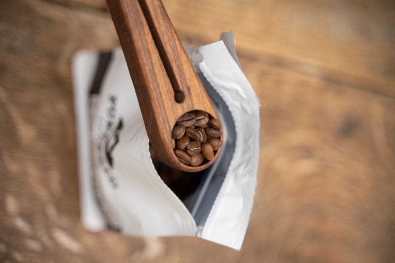 May include: A wooden scoop filled with roasted coffee beans, being poured into a white bag with a black label.