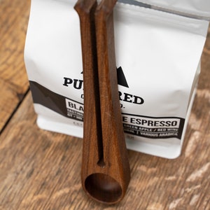 May include: A wooden scoop with a handle that is shaped like a pair of tongs. The scoop is holding a bag of coffee beans that says "Pured Co. Black Espresso" on the front.