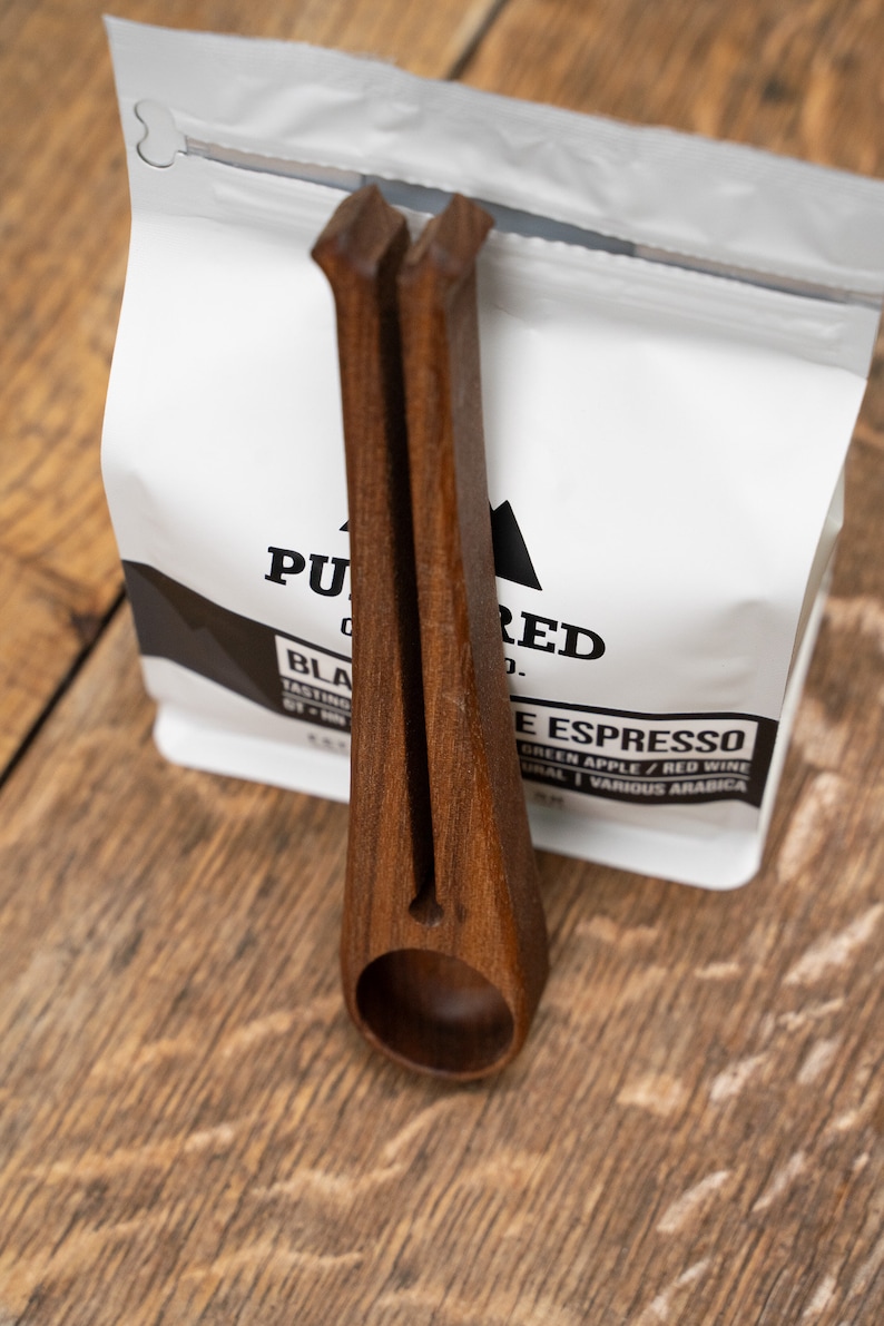 May include: A wooden scoop with a handle that is shaped like a pair of tongs. The scoop is holding a bag of coffee beans that says "Pured Co. Black Espresso" on the front.