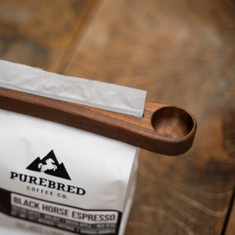 May include: A wooden coffee scoop with a handle and a round scoop. The scoop is attached to a white coffee bag with a black logo that says "Purebred Coffee Co. Black Horse Espresso".