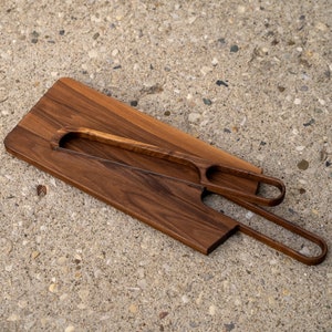 May include: A wooden serving board with two integrated serving tools. The board is made of dark wood and has a natural finish. The tools are made of the same wood and have a smooth, rounded shape.