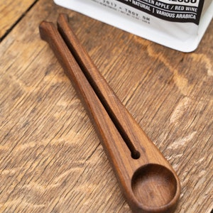 May include: A wooden coffee scoop with a handle and a rounded scoop at the end. The scoop is made of dark wood and has a natural finish. The scoop is sitting on a wooden surface.