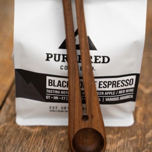 May include: Two wooden coffee scoops with a white bag of coffee beans in the background. The bag has the text "PUR 1 RED CO" and "BLACK DESPRESSO" printed on it.