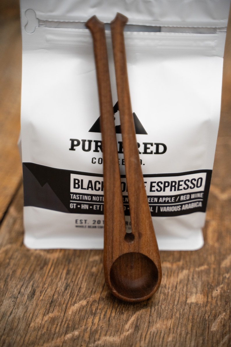 May include: Two wooden coffee scoops with a white bag of coffee beans in the background. The bag has the text "PUR 1 RED CO" and "BLACK DESPRESSO" printed on it.