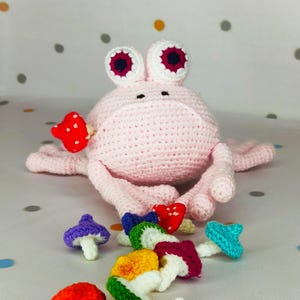Puede incluir: Una rana de ganchillo rosa claro hecha a mano con ojos grandes con borde blanco y una colección de coloridas setas de ganchillo. La rana tiene un diseño caprichoso, con una seta en la boca. Las setas son rojas, moradas, azules, verdes, amarillas y granates.