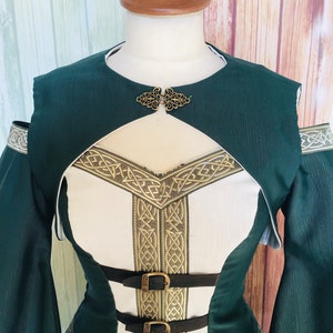 May include: A green and white medieval-style dress with gold trim and black leather straps. The dress has a high neckline, long sleeves, and a decorative clasp. The bodice features a white panel with gold Celtic knotwork. The dress is on a mannequin.