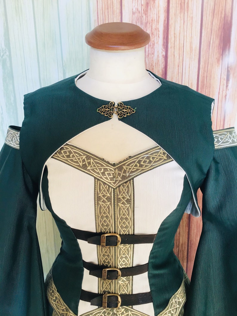 May include: A green and white medieval-style dress with gold trim and black leather straps. The dress has a high neckline, long sleeves, and a decorative clasp. The bodice features a white panel with gold Celtic knotwork. The dress is on a mannequin.