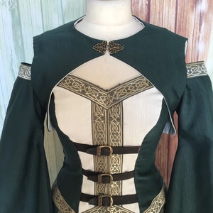 May include: A green and white medieval-style dress with gold trim and three brown leather straps. The dress has long sleeves and a decorative clasp at the neckline. The bodice is white with gold Celtic knotwork.