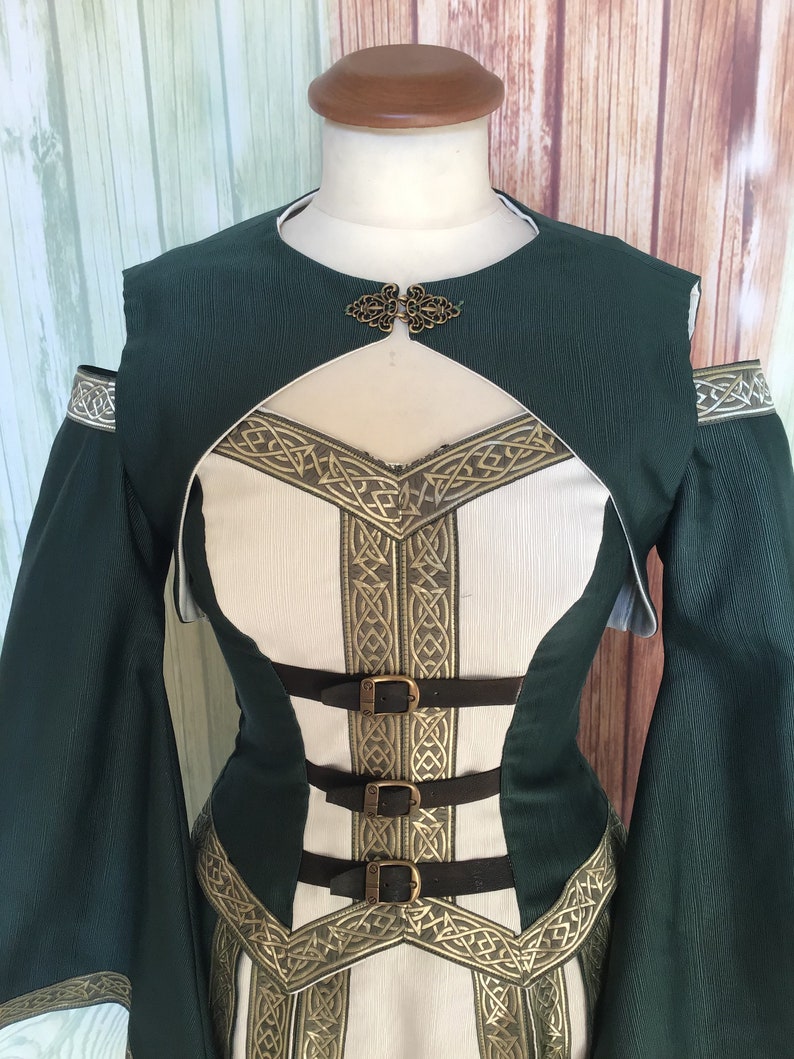 May include: A green and white medieval-style dress with gold trim and three brown leather straps. The dress has long sleeves and a decorative clasp at the neckline. The bodice is white with gold Celtic knotwork.