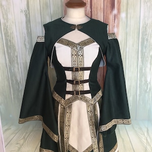 May include: A green and white medieval-style dress with long, flowing sleeves. The dress features gold trim and accents, with a decorative clasp at the neckline and brown belt details. The dress is displayed on a mannequin.