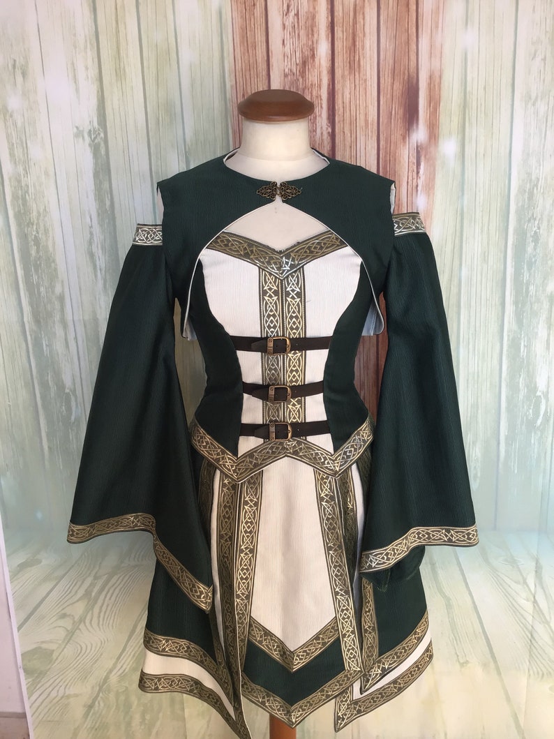 May include: A green and white medieval-style dress with long, flowing sleeves. The dress features gold trim and accents, with a decorative clasp at the neckline and brown belt details. The dress is displayed on a mannequin.