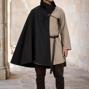 May include: A two-toned garment featuring a black cape attached to a beige tunic with a high collar. The tunic is secured with a dark brown leather belt at the waist. The ensemble is paired with dark brown trousers and dark brown leather boots.