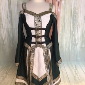 May include: A green and white medieval-style dress with gold trim and accents. The dress features long sleeves, a fitted bodice with brown straps, and a flared skirt. The off-the-shoulder design and intricate detailing give it a regal appearance.