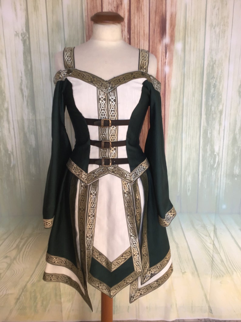 May include: A green and white medieval-style dress with gold trim and accents. The dress features long sleeves, a fitted bodice with brown straps, and a flared skirt. The off-the-shoulder design and intricate detailing give it a regal appearance.