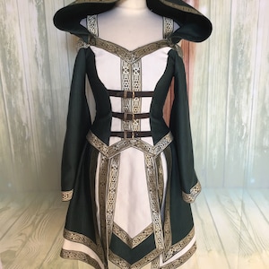 May include: A green and white hooded dress with gold trim and brown buckles. The dress has long sleeves and a detailed design around the neckline, waist, and hem. The dress is displayed on a mannequin.