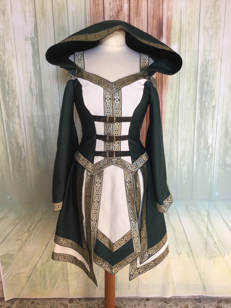 May include: A green and white hooded dress with gold trim and brown buckles. The dress has long sleeves and a detailed design around the neckline, waist, and hem. The dress is displayed on a mannequin.