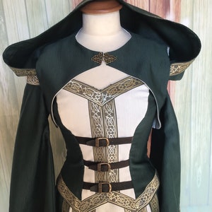May include: A green and white medieval-style dress with a hood. The dress has a bodice with brown straps and buckles, and gold-coloured trim with a Celtic knot design. The sleeves are long and flared.