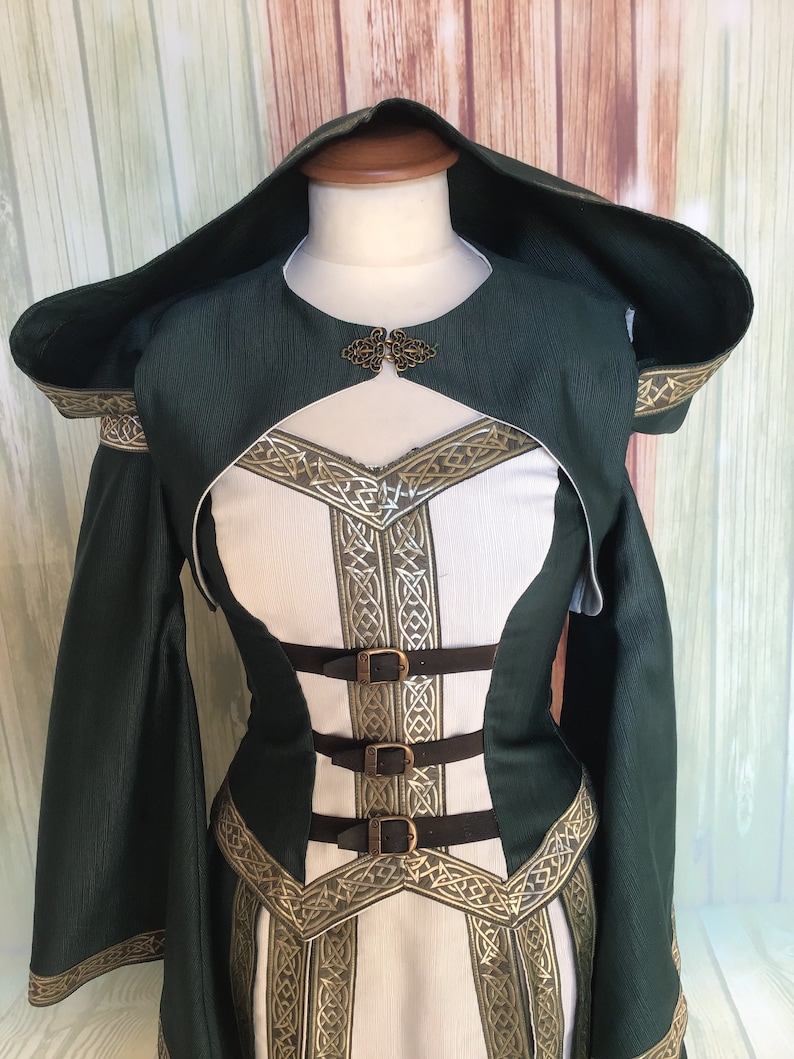 May include: A green and white medieval-style dress with a hood. The dress has a bodice with brown straps and buckles, and gold-coloured trim with a Celtic knot design. The sleeves are long and flared.