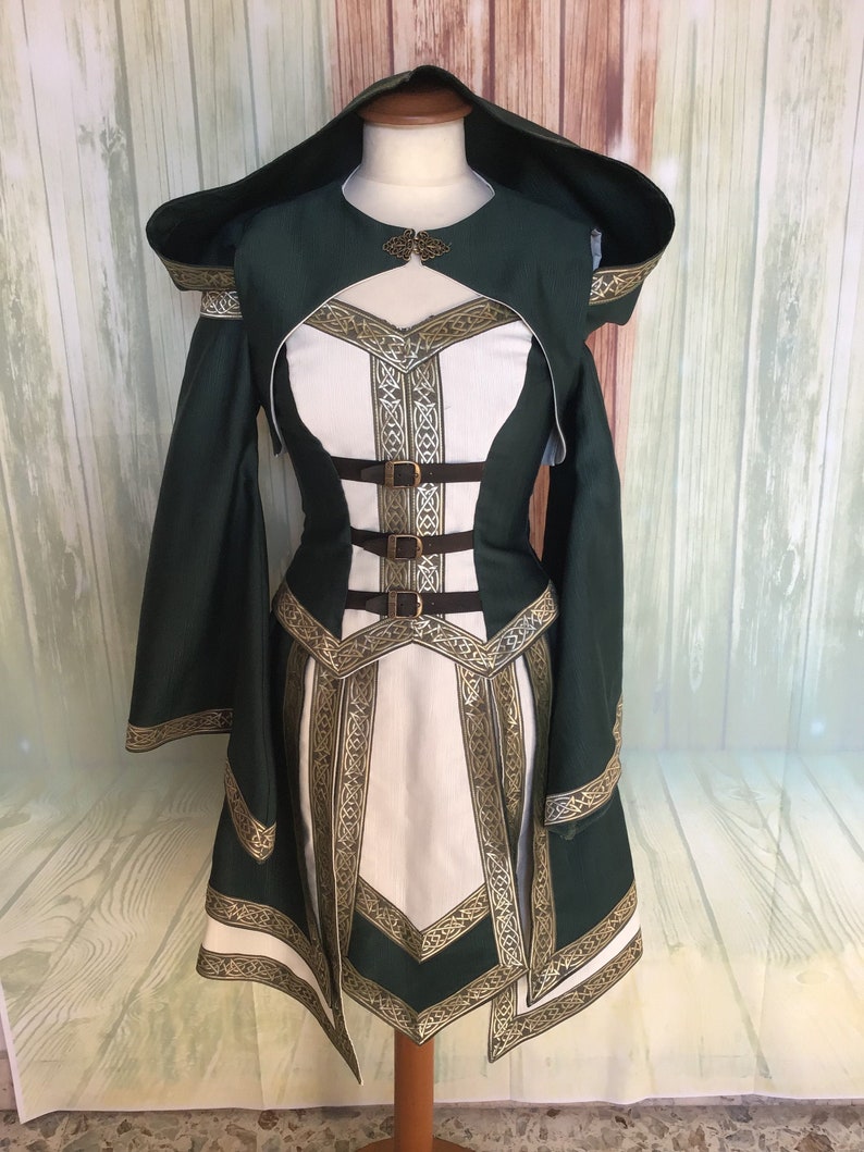 May include: A green and white medieval-style dress with a hood, displayed on a mannequin. The dress features gold trim and brown leather straps. The design includes intricate patterns and a decorative clasp at the neckline.
