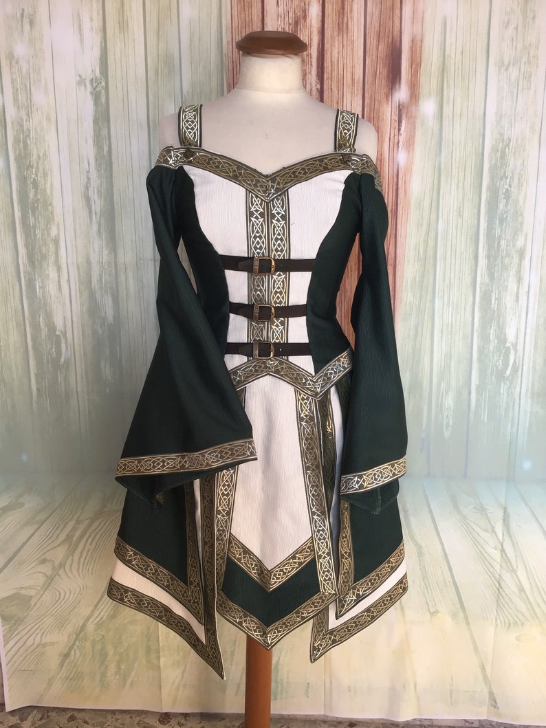 May include: A green and white medieval-style dress with gold trim and brown buckles. The dress has off-the-shoulder sleeves and a fitted bodice. The design includes intricate gold patterns along the edges and straps, creating a historical aesthetic.